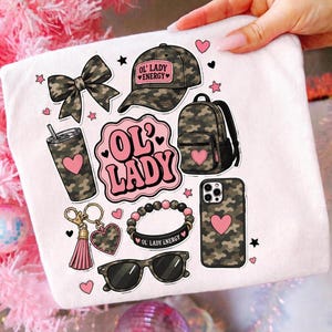 Könnte beinhalten: Ein weißes T-Shirt mit einem Grafikdruck von camouflagemusterten Gegenständen, darunter eine Baseballkappe mit dem Text OL' LADY ENERGY, ein Rucksack, ein Becher, ein Schlüsselanhänger, ein Armband, eine Sonnenbrille, eine Handyhülle und eine Schleife.
