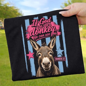 Könnte beinhalten: Ein schwarzes T-Shirt mit einer Esel-Grafik und dem Text "If God can use a donkey, He can use you." Das Design enthält hellblaue vertikale Streifen, einen rosa Stern, einen hellblauen Stern und ein kleines rechteckiges Feld mit der Aufschrift "NUMBERS 22."