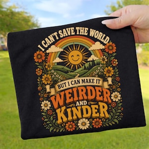 May include: A black t-shirt featuring a graphic print of a smiling sun, a rainbow, and rolling hills surrounded by orange and white flowers. The text reads, I CAN'T SAVE THE WORLD BUT I CAN MAKE IT WEIRDER AND KINDER in bold, colorful lettering.