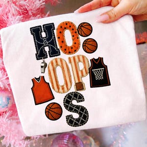 Könnte beinhalten: Ein weißes T-Shirt mit dem Wort HOOPS in großen, dekorativen Buchstaben mit Mustern. Das H ist dunkelblau, das O ist orange mit schwarzen Punkten, das O ist orange-weiß gestreift, das P ist orange-weiß gestreift und das S ist schwarz mit einem weißen Netzmuster. Das Design enthält zwei Basketbälle, zwei Basketballtrikots und eine silberne Pfeife.
