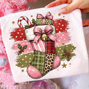 May include: A white t-shirt featuring a graphic of a patchwork Christmas stocking with pink, green, and red patterns, including plaid and leopard print. The stocking is adorned with a pink bow, a gold bell, a candy cane, and holly berries.