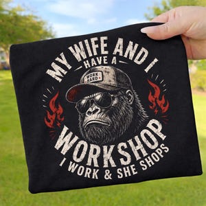 Könnte beinhalten: Ein schwarzes T-Shirt mit einer Grafik eines Gorillas, der eine Sonnenbrille und eine Kappe mit der Aufschrift WORK HARD trägt. Der Text lautet MY WIFE AND I HAVE A WORKSHOP I WORK & SHE SHOPS, umrahmt von roten Flammen in weißer Schrift.