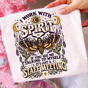 Könnte beinhalten: Ein weißes T-Shirt mit einem Grafikdruck eines Nachtfalters, der ein Totenkopf- und Augenmuster auf dem Körper trägt. Das Design umfasst Mondsicheln, Kristalle und Lavendelzweige mit dem Text: I WORK WITH SPIRIT IF YOU SEE ME TALKING TO MYSELF, IT'S JUST A STAFF MEETING.