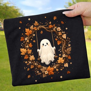 Könnte beinhalten: Ein schwarzes T-Shirt mit einer Grafik eines weißen Geistes, der auf einer Holzschaukel sitzt. Die Schaukel hängt in einem kreisförmigen Kranz aus braunen Ästen, orangefarbenen und gelben Herbstblättern sowie kleinen weißen Blüten.