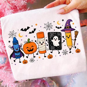 Könnte beinhalten: Ein weißes T-Shirt mit einer bunten Grafik von Cartoon-Schulmaterialien, die für Halloween verkleidet sind. Das Design zeigt einen blauen Wachsmalstift als Vampir, einen Kürbis, einen Klebestift als Mumie, ein Gespenst im Buch und einen Bleistift mit Hexenhut.