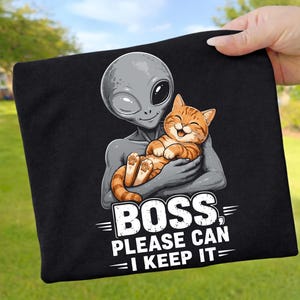 Könnte beinhalten: Schwarzes T-Shirt mit einem Cartoon-Alien, das eine lächelnde orangefarbene Tabby-Katze hält. Der Text "BOSS, PLEASE CAN I KEEP IT?" ist weiß gedruckt. Das Design befindet sich vor einem grünen Hintergrund.