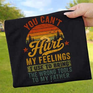 Könnte beinhalten: Ein schwarzes T-Shirt mit einer Grafik, die einen Sonnenuntergang, Kiefern und den Text zeigt: YOU CAN'T Hurt MY FEELINGS I USE TO BRING THE WRONG TOOLS TO MY FATHER. Das Design verwendet eine abgenutzte Vintage-Schrift in Orange, Gelb und Grün.