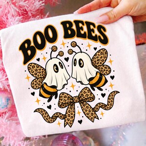 Könnte beinhalten: Ein weißes T-Shirt mit einer Grafik von zwei Bienen in weißen Geisterkostümen. Das Design zeigt den schwarzen Schriftzug BOO BEES, eine Schleife mit Leopardenmuster sowie kleine gelbe Sterne und schwarze Herzen auf dem Stoff verteilt.