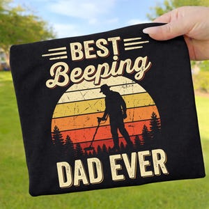 Könnte beinhalten: Ein schwarzes T-Shirt mit einem Grafikdruck einer Person, die einen Metalldetektor benutzt. Das Design zeigt einen Sonnenuntergang mit Tannen und den Text BEST BEEPING DAD EVER in einer abgenutzten Retro-Schriftart in Gelb- und Orangetönen.