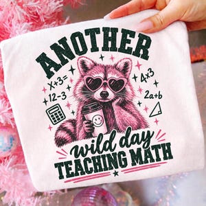 Könnte beinhalten: Ein weißes T-Shirt mit einer Grafik eines rosa Waschbären, der herzförmige Sonnenbrillen trägt und einen Kaffeebecher hält. Der Text lautet "ANOTHER wild day TEACHING MATH", umgeben von Mathegleichungen, einem Taschenrechner und einem Dreieck.