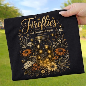 Könnte beinhalten: Ein schwarzes T-Shirt mit einer goldenen Grafik eines Einmachglases voller leuchtender Glühwürmchen, umgeben von Wildblumen. Der Text Fireflies and front porch nights steht in einer dekorativen Schrift über dem Glas.