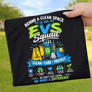 May include: A black t-shirt featuring a graphic with the text BEHIND A CLEAN SPACE IS AN EVS SQUAD. The design includes a cleaning cart, a yellow caution sign, a blue bucket, and icons representing safety, sustainability, respect, and teamwork.