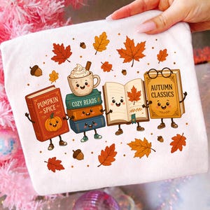 Könnte beinhalten: Ein weißes T-Shirt mit einer Grafik von vier anthropomorphen Büchern und einer Tasse Kakao. Die Bücher sind mit Pumpkin Spice, Cozy Reads und Autumn Classics beschriftet. Das Design enthält orangefarbene Herbstblätter und Eicheln.