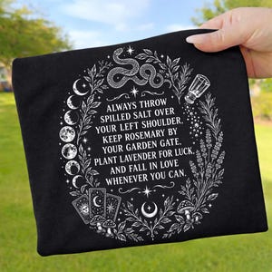 Könnte beinhalten: Ein schwarzes T-Shirt mit einem weißen Grafikdruck von einer Schlange, Mondphasen, Tarotkarten, Pilzen und Lavendel. Der Text lautet: ALWAYS THROW SPILLED SALT OVER YOUR LEFT SHOULDER, KEEP ROSEMARY BY YOUR GARDEN GATE, PLANT LAVENDER FOR LUCK, AND FALL IN LOVE WHENEVER YOU CAN.