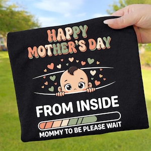 May include: Black t-shirt with the text "HAPPY MOTHER'S DAY" in orange and green. A baby graphic peeks out from a white line, with the words "FROM INSIDE" and "MOMMY TO BE PLEASE WAIT" below. Small heart shapes are scattered around the baby.
