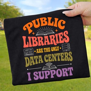 Könnte beinhalten: Ein schwarzes T-Shirt mit dem orangefarbenen Text PUBLIC LIBRARIES, rotem Text ARE THE ONLY, goldenem Text DATA CENTERS und violettem Text I SUPPORT. Das Design enthält ein weißes Strichzeichnungs-Buchsymbol, eine Bibliothekskarte, einen Server und ein WLAN-Buch.