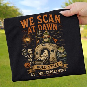 May include: A black t-shirt featuring a graphic print with the text WE SCAN AT DAWN, HOLD STILL, and CT MRI DEPARTMENT. The design shows frogs, a skeleton, and a ghost around an MRI machine, with a pumpkin and a lantern in orange and green tones.