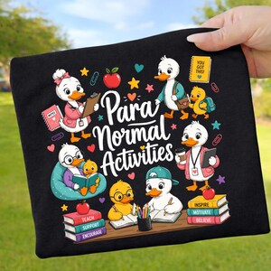 Könnte beinhalten: Ein schwarzer Stoffartikel mit einer bunten Grafik von Zeichentrickenten in einem Klassenzimmer und dem Text "Para Normal Activities". Das Design zeigt Enten mit Büchern, Äpfeln, Stiften und Bücherstapeln mit den Aufschriften "Teach", "Support", "Encourage", "Inspire", "Motivate", "Believe".