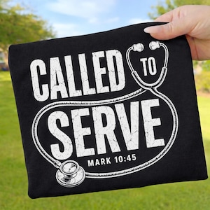 May include: A black t-shirt featuring white distressed text that reads CALLED TO SERVE with the reference MARK 10:45. The design incorporates a white graphic of a stethoscope that frames the text, creating a medical-themed visual element.