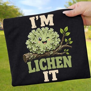 Könnte beinhalten: Schwarzes T-Shirt mit einem grünen Cartoon-Flechten-Charakter, der zwinkert. Der Text "I'M LICHEN IT" ist über und unter der Flechten-Illustration aufgedruckt. Das Design beinhaltet einen Ast mit Blättern.