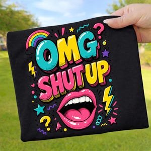 Könnte beinhalten: Schwarzes T-Shirt mit einem farbenfrohen Grafikdesign. Das Design zeigt die Worte "OMG SHUTUP" in fetten, umrandeten Buchstaben, einen Regenbogen, einen offenen Mund, Sterne und Blitze. Das T-Shirt wird von einer Hand gehalten.