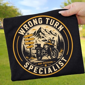 May include: A black t-shirt featuring a circular graphic with a yellow border. The design shows a motorcycle parked near a wooden signpost in front of mountains and trees. The text reads WRONG TURN SPECIALIST and ADV DIVISION in yellow.