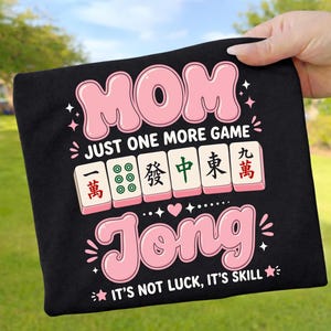May include: A black t-shirt featuring a graphic with the pink text MOM, the white text JUST ONE MORE GAME, and the pink text JONG. Between the text are six mahjong tiles with Chinese characters and green dots. The bottom text reads IT'S NOT LUCK, IT'S SKILL.