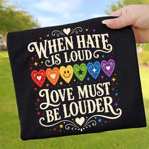 Könnte beinhalten: Ein schwarzes T-Shirt mit dem weißen Text "WHEN HATE IS LOUD LOVE MUST BE LOUDER" und einer Reihe von sechs bunten Herzen in Rot, Orange, Gelb, Grün, Blau und Lila, akzentuiert mit kleinen Sternen und dekorativen Verzierungen.