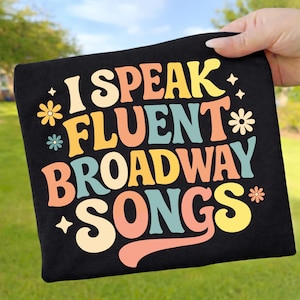 May include: Black t-shirt with the text "I Speak Fluent Broadway Songs" in colorful, retro-style lettering. The design includes flowers and stars. The shirt is held up against a background of green grass and a blue sky.