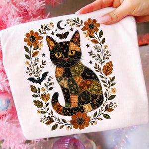Könnte beinhalten: Ein weißes T-Shirt mit einem Grafikdruck einer Katze im Patchwork-Stil mit gelben Augen, umgeben von einem Kranz aus herbstfarbenen Blumen, Blättern, einer Mondsichel, Sternen, einem Schmetterling und einer kleinen Fledermaussilhouette in Orange- und Brauntönen.