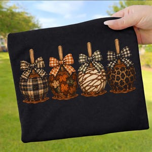 Könnte beinhalten: Ein schwarzes T-Shirt mit einer Grafik von vier kandierten Äpfeln in einer Reihe. Jeder Apfel hat einen Holzstiel und eine dekorative Schleife. Die Designs umfassen schwarzes Karomuster, orangefarbene Herbstblätter, weiße Glasur und Leopardenmuster.