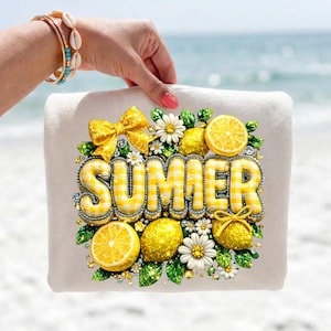 Könnte beinhalten: Ein weißes Strandtuch mit dem Wort "SUMMER" in gelben Vichy-Buchstaben. Das Tuch ist mit Zitronen, Gänseblümchen und gelben Schleifen verziert. Der Hintergrund ist ein verschwommenes Bild des Ozeans.