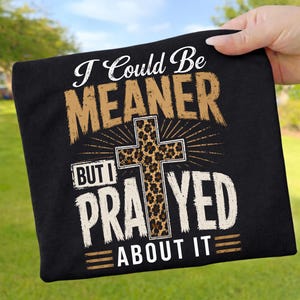 Könnte beinhalten: Ein schwarzes T-Shirt mit dem weißen und beigen Text I COULD BE MEANER BUT I PRAYED ABOUT IT. Das Design zeigt ein zentrales Kreuz mit braun-schwarzem Leopardenmuster, umgeben von strahlenförmigen Linien auf einem dunklen Stoffhintergrund.