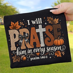 Könnte beinhalten: Ein schwarzes T-Shirt mit dem Text I will PRAISE him in every season und psalm 146:2. Das Design zeigt ein weißes Kreuz, eine schwarze Schleife, einen orangefarbenen Kürbis, eine Eichel und braune Herbstblätter mit kleinen weißen Sternen auf Schwarz.