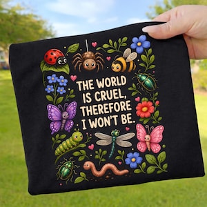 Könnte beinhalten: Ein schwarzer Stoffartikel mit einer farbenfrohen Illustration verschiedener Insekten und Blumen, die den Text "THE WORLD IS CRUEL, THEREFORE I WON'T BE" umgeben. Das Design zeigt einen Marienkäfer, eine Spinne, eine Biene, Schmetterlinge und eine Libelle.