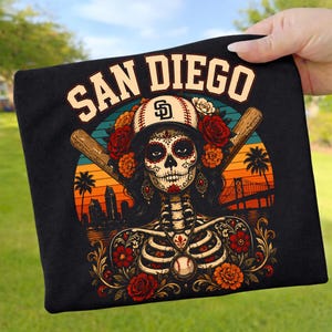 May include: Black t-shirt featuring a sugar skull design with a baseball cap and the text "SAN DIEGO". The design includes baseball bats, flowers, and a cityscape with a bridge.