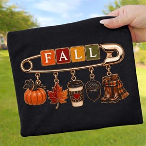 Könnte beinhalten: Ein schwarzes T-Shirt mit einer Grafik einer großen goldenen Sicherheitsnadel, auf der das Wort FALL in bunten quadratischen Blöcken steht. An der Nadel hängen Anhänger in Form eines orangefarbenen Kürbisses, eines roten Ahornblatts, eines Kaffeebechers, einer Eichel und brauner Stiefel.