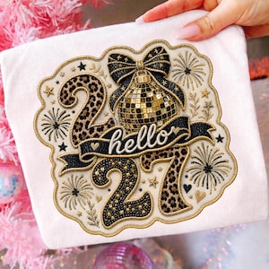 Puede incluir: Un parche de tela blanca con un diseño bordado que incluye el texto hello, los números 2027, una bola de discoteca y un lazo. Los números están decorados con estampado de leopardo y lentejuelas negras, rodeados de fuegos artificiales y estrellas.