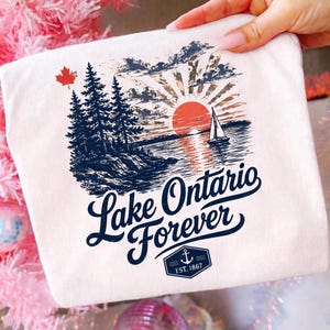 May include: A white t-shirt featuring a graphic of a sunset over Lake Ontario with a sailboat, pine trees, and a red maple leaf. The text reads Lake Ontario Forever with an anchor emblem and the text EST. 1867 below the main graphic design.