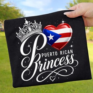 Könnte beinhalten: Schwarzes T-Shirt mit einem weißen, kursiven "Princess"-Design. Das Design umfasst eine Krone, ein Herz mit der puerto-ricanischen Flagge und die Worte "Puerto Rican". Das T-Shirt wird vor einem grünen Hintergrund gehalten.