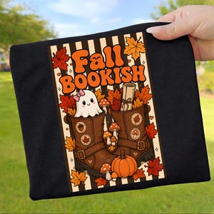Könnte beinhalten: Ein schwarzes T-Shirt mit einem Grafikdruck, der den Text "Fall BOOKISH" in orangefarbenen Buchstaben zeigt. Das Design umfasst ein weißes Gespenst mit einer rosa Schleife, braune Stiefel, einen Kürbis, Pilze und orangefarbene Ahornblätter.