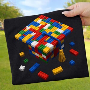 Könnte beinhalten: Ein schwarzes Design für eine Abschlusskappe aus bunten Spielzeugbausteinen. Die Kappe hat eine blaue Kordel und eine goldene Quaste. Einzelne Spielzeugbausteine in Rot, Gelb, Blau, Grün und Weiß sind um die Kappe herum verstreut.