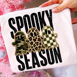 Könnte beinhalten: Ein weißes T-Shirt mit dem schwarzen, abgenutzten Schriftzug SPOOKY SEASON und drei geisterförmigen Aufnähern. Die Geister haben goldfarbene, gestickte Ränder und zeigen ein grünes Tarnmuster, ein braunes Leopardenmuster und ein grün-weißes Karomuster.