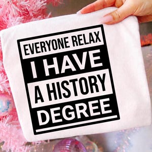 Op de afbeelding: Een wit t-shirt met een opvallende zwarte rechthoekige afbeelding met de tekst "EVERYONE RELAX I HAVE A HISTORY DEGREE" gedrukt in witte en zwarte blokletters. Het ontwerp is gecentreerd op de voorkant van het kledingstuk met strakke lijnen.