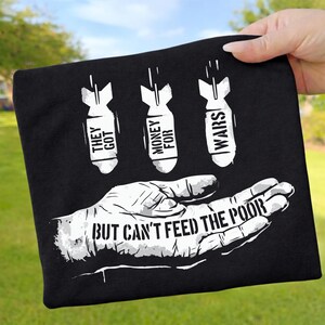Könnte beinhalten: Schwarzes T-Shirt mit weißer Grafik. Die Grafik zeigt drei stilisierte Bomben mit den Worten "THEY GOT", "MONEY FOR" und "WARS". Darunter eine Hand mit dem Text "BUT CAN'T FEED THE POOR".