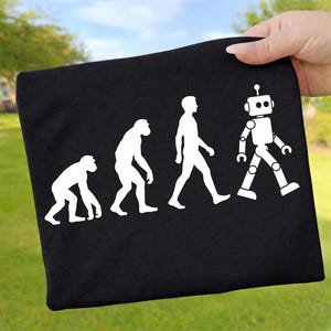 Pode incluir: Uma t-shirt preta com uma impressão gráfica branca que retrata a progressão evolutiva de um primata para um humano, terminando com um robô. O design mostra quatro figuras em linha, ilustrando a transição de macaco para máquina.