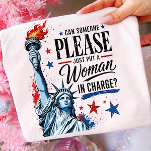 Könnte beinhalten: Ein weißes T-Shirt mit einer Grafik der Freiheitsstatue, die eine Fackel mit orangefarbenen Flammen hält. Der Text lautet: CAN SOMEONE PLEASE JUST PUT A WOMAN IN CHARGE? Das Design enthält rote und blaue Sterne sowie Farbspritzer-Akzente.
