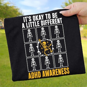 Pode incluir: Uma t-shirt preta com o texto branco IT'S OKAY TO BE A LITTLE DIFFERENT e ADHD AWARENESS. O design inclui uma grelha de oito esqueletos brancos e um esqueleto amarelo central a dançar dentro de um recorte circular irregular.