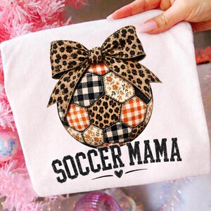 Könnte beinhalten: Ein weißes T-Shirt mit einer Grafik eines Fußballs, der mit einem Patchwork-Muster aus Leopardenmuster, schwarz-weißem Karomuster und orangefarbenen Blumen verziert ist, gekrönt von einer großen Leoparden-Schleife und dem Text SOCCER MAMA.