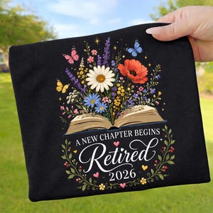 Könnte beinhalten: Schwarzes T-Shirt mit einem floralen Design mit Schmetterlingen, Gänseblümchen und Mohnblumen über einem offenen Buch. Der Text lautet "A New Chapter Begins Retired 2026" in einer dekorativen Schriftart, umgeben von einem floralen Kranz.